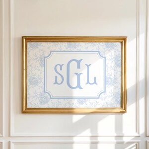 May include: A framed monogram art piece featuring the letters "SGL" in light blue. The artwork is set against a white background with a light blue floral pattern and framed in a gold-colored frame. The artwork is hanging on a white wall.