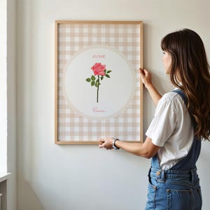 May include: A framed print featuring a pink rose illustration. The artwork is set against a beige and white gingham pattern, with the word "June" and "Rose" displayed. The frame is made of light-colored wood.