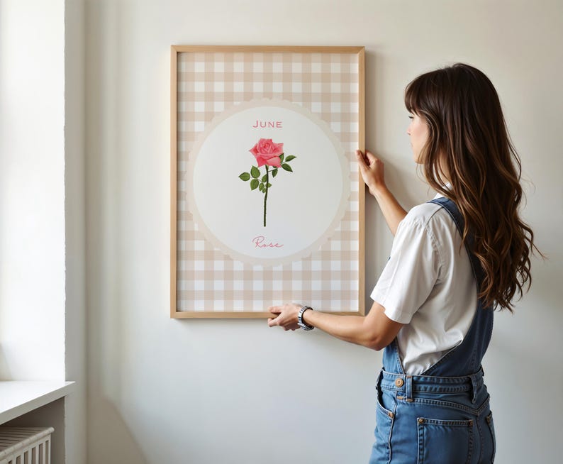 May include: A framed print featuring a pink rose illustration. The artwork is set against a beige and white gingham pattern, with the word "June" and "Rose" displayed. The frame is made of light-colored wood.