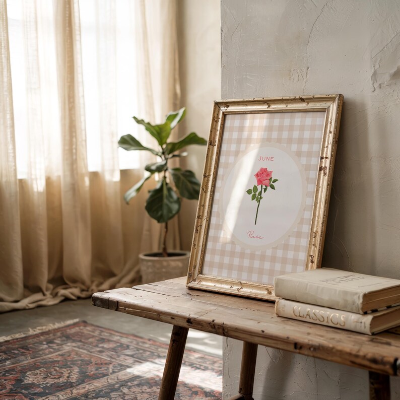 May include: A framed print featuring a pink rose illustration with the text "June" and "Rose" on a beige gingham background. The gold frame rests on a wooden bench with two books. A potted plant and window with beige curtains are in the background.