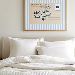 May include: A framed postcard print with a light blue and white striped background. The postcard reads "Meet me in State College!" in navy blue script, with a heart and paw print graphic. The frame is a light brown color. The print hangs above a bed with white bedding.
