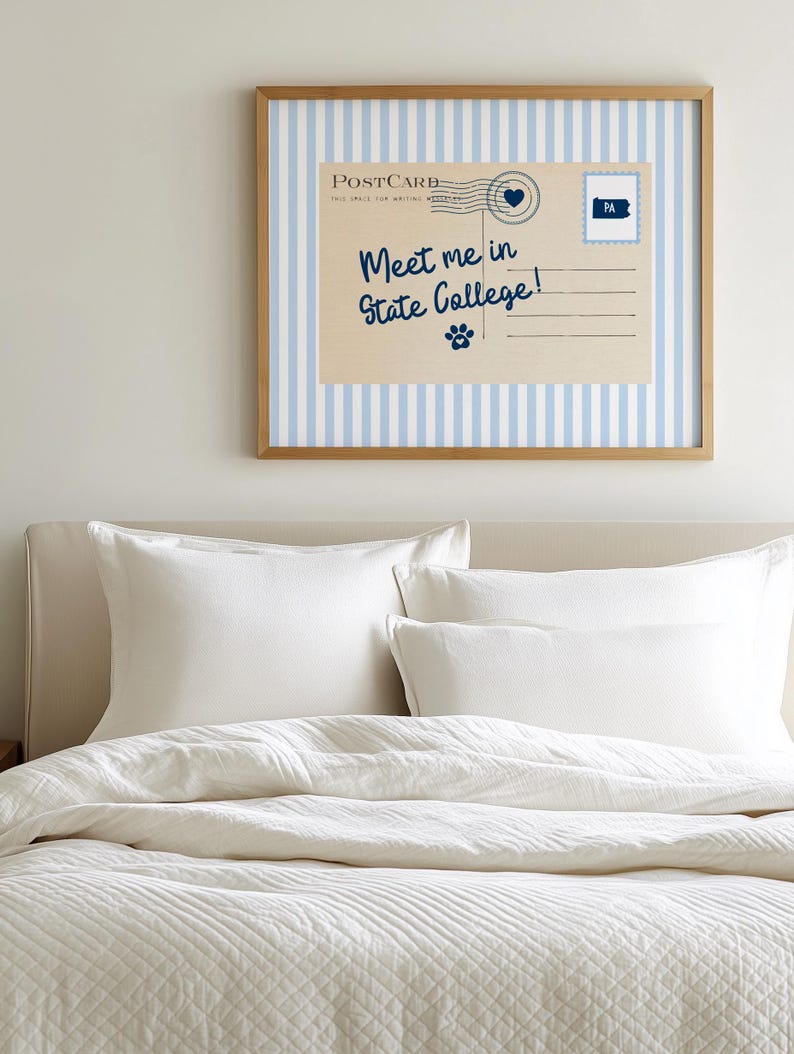 May include: A framed postcard print with a light blue and white striped background. The postcard reads "Meet me in State College!" in navy blue script, with a heart and paw print graphic. The frame is a light brown color. The print hangs above a bed with white bedding.