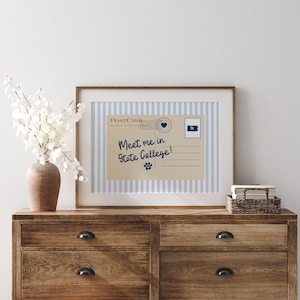 May include: A framed postcard print with the text "Meet me in State College!" on a striped blue and white background. The postcard features a heart stamp and a Pennsylvania state outline. The print is displayed on a wooden dresser.