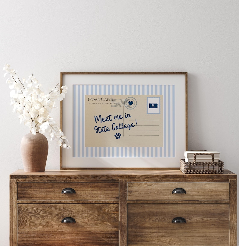 May include: A framed postcard print with the text "Meet me in State College!" on a striped blue and white background. The postcard features a heart stamp and a Pennsylvania state outline. The print is displayed on a wooden dresser.
