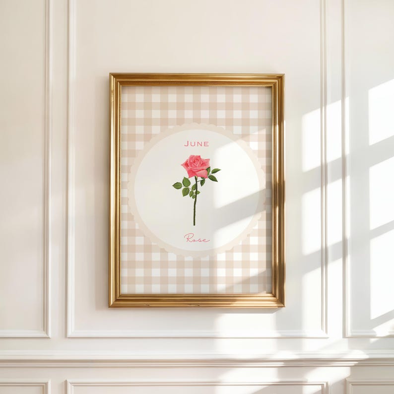 May include: A framed print featuring a pink rose illustration. The artwork is set against a beige gingham pattern and a cream-colored circle. The word "June" is at the top, and "Rose" is at the bottom. The frame is gold.