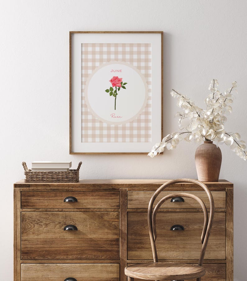 May include: A framed print featuring a pink rose illustration. The artwork is set against a beige and white gingham pattern, with the words "JUNE" and "Rose" displayed. The frame is brown, and the print is displayed on a wooden dresser.