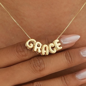 Könnte beinhalten: Eine goldfarbene Halskette mit dem Namen "GRACE" in einzelnen, abgerundeten Buchstaben. Die Halskette wird um den Hals getragen, wobei die Buchstaben zentriert sind. Die Kette ist filigran und die Buchstaben sind verbunden. Der Hintergrund ist die Haut einer Person.