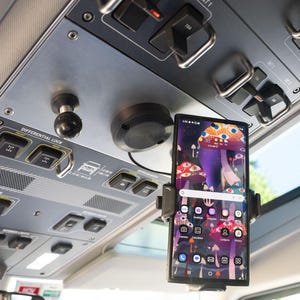 May include: A smartphone mounted to the overhead console of a vehicle. The console features various black toggle switches, buttons, and labels including Differential Lock. The phone screen displays a colorful mushroom-themed wallpaper and app icons.