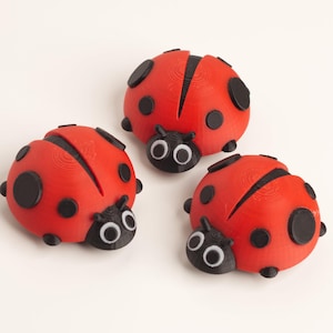 May include: Three red ladybug figurines with black spots and black heads with white-eyed details are arranged on a white surface. The ladybugs have a rounded shape and are a decorative item.
