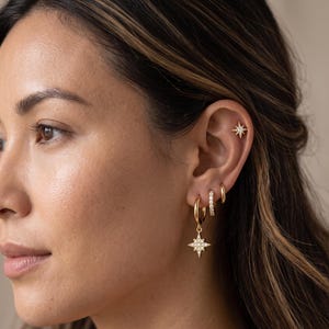 Puede incluir: Un conjunto de pendientes dorados que consta de un pequeño perno en forma de estrella en la parte superior de la oreja, dos aros dorados sencillos y un aro más grande con un dije colgante en forma de estrella con pequeñas piedras claras.