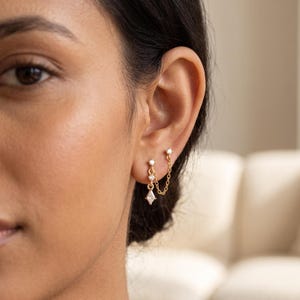 May include: A gold chain earring featuring two studs connected by a delicate chain. The earring includes three small clear stones, with one stone set in a kite-shaped pendant hanging from the chain. The design spans two ear piercings.