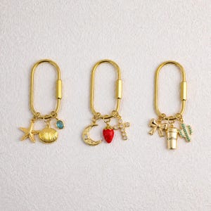 Peut inclure: Trois porte-clés mousquetons ovales dorés. Le premier comporte une étoile de mer, un coquillage et une breloque en pierre bleue. Le deuxième a un croissant de lune, une fraise rouge et une croix en cristal. Le troisième inclut un nœud, une tasse de café et un cactus.