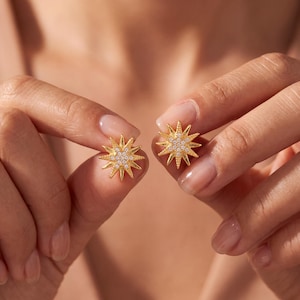 May include: A pair of gold starburst stud earrings featuring a textured metallic finish and a cluster of small, clear, round-cut stones embedded in the center of each star, radiating outward with pointed rays in a symmetrical design.