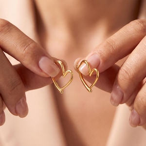 May include: A pair of gold-toned hoop earrings shaped like open hearts. The earrings feature a smooth, polished metallic finish with a continuous wire design that forms a heart silhouette, held between fingers to display the curved structure.