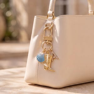May include: A cream-colored handbag featuring a gold-toned metal keychain attached to the handle. The keychain includes a light blue seashell charm, a gold-toned cowboy boot charm with intricate detailing, and a letter A charm with small crystals.