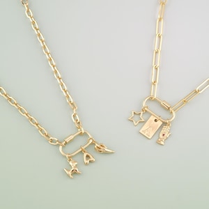 Puede incluir: Dos collares dorados con colgantes de abalorios. Un collar presenta un perro, un corazón y un cuerno. El otro collar tiene una estrella, una etiqueta con la letra "A" y una mano. Los collares tienen un diseño de eslabones de cadena.