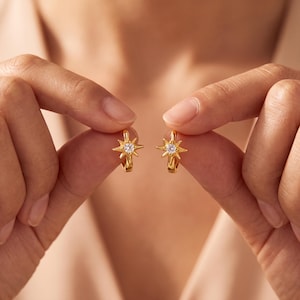May include: A pair of gold hoop earrings featuring a starburst design. Each earring is centered with a single clear, round-cut gemstone. The starburst motif has eight pointed rays radiating from the central stone, creating a celestial look.
