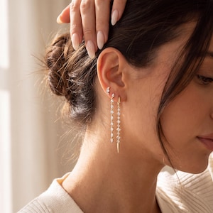 May include: A set of three gold-toned earrings worn in one ear. The top earring is a small stud, the middle is a long chain with marquise-cut stones, and the bottom is a hoop earring with a vertical chain of round stones ending in a gold spike.