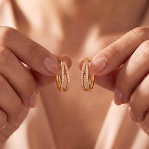 May include: A pair of gold hoop earrings featuring a double-band design. Each hoop is accented with a row of small, clear, sparkling stones embedded along the front surface of the bands, creating a refined and polished appearance for these earrings.