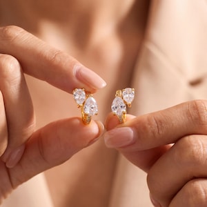 May include: A pair of gold hoop earrings featuring two clear pear-shaped stones arranged in a cluster. Each earring has a small gold hoop base with a polished finish, supporting a larger teardrop-shaped stone and a smaller round stone above it.