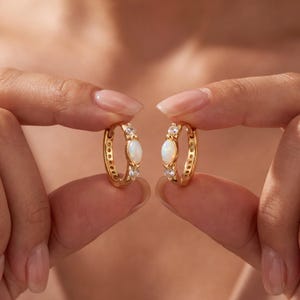 May include: A pair of gold hoop earrings, each featuring a central marquise-cut opal stone flanked by two small round clear stones. The hoops have a polished gold finish and a hinged closure mechanism, showcasing a delicate and refined design.