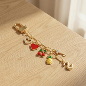 Pode incluir: Um porta-chaves de metal dourado com fecho lagosta e uma corrente com vários berloques: um coração com cristais, um coração vermelho sólido, um par de cerejas vermelhas com folhas verdes, um abacaxi amarelo, uma chave de cristal e a letra S.