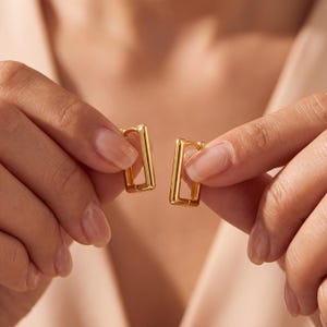 May include: A pair of gold-toned hoop earrings featuring a rectangular shape with rounded edges. The earrings have a polished, reflective surface and a hinged closure mechanism, showcasing a clean, geometric design that is simple and uniform.