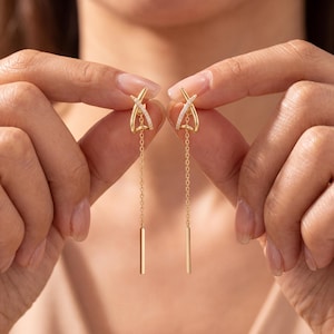 May include: A pair of gold drop earrings featuring a delicate chain design. Each earring has a top section with a crisscross shape accented by small clear stones, connected to a thin gold chain that ends in a smooth, straight metal bar.
