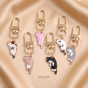 Può includere: Sei portachiavi in metallo dorato con ciondoli smaltati a forma di gatto in vari colori, tra cui calico, rosa, grigio, bianco e nero, e crema. Ogni ciondolo è fissato a un moschettone tramite un anellino. Il testo HAND MADE è stampato in basso.