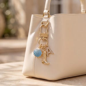 May include: A cream-coloured handbag with gold hardware and a charm cluster. The charms include a blue seashell, a gold cowboy boot, and a letter "A" encrusted with rhinestones. The bag is made of smooth leather.