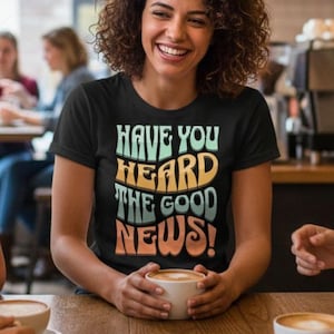 May include: A group of friends at a cafe, laughing and enjoying coffee. A woman in the center wears a black t-shirt with the text "HAVE YOU HEARD THE GOOD NEWS!" in retro colors. The scene is warm and inviting, with a wooden table and cafe setting.