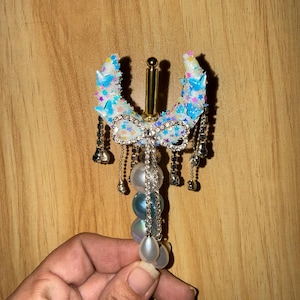 May include: A decorative hair accessory featuring a U-shaped design with blue star accents and a rhinestone bow. Dangling chains with small silver bells and a strand of iridescent beads complete the design.