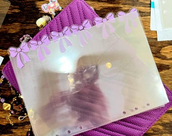 Può includere: Una tasca per planner trasparente e iridescente con una fila di fiocchi viola chiaro lungo il bordo superiore. La tasca è appoggiata su una copertina per planner viola scuro strutturata. Sono visibili gioielli decorativi e un ciondolo.