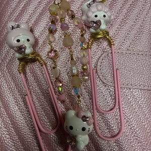 May include: Two large pink paper clips decorated with gold wire and small white cartoon character figurines holding pink heart-shaped gems. A central beaded chain connects the clips, featuring translucent pink, white, and iridescent beads.