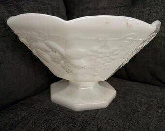 May include: A white, vintage-style pedestal bowl with a scalloped rim. The bowl features a detailed embossed grape and leaf design on the sides. The bowl sits on a hexagonal base, creating a classic and elegant look.