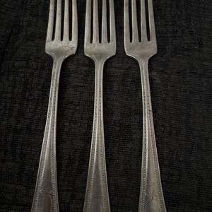 May include: Three vintage silver forks with tines and ornate handles. The forks are arranged in a row against a dark, textured background. The handles have a decorative design near the base.