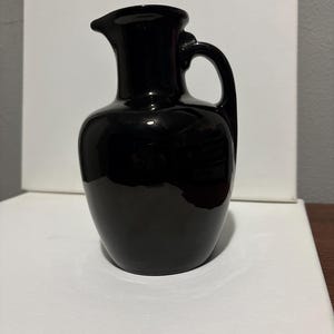 May include: A glossy black ceramic pitcher with a handle and a spout. The pitcher has a rounded body and a smooth, reflective surface. The handle is arched, and the spout is slightly curved.
