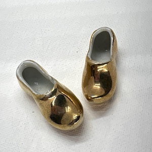 May include: Two miniature gold-colored clogs with white interiors. The clogs are made of a shiny material, reflecting light. The clogs are side by side, suggesting they are a pair. The clogs are likely decorative items or collectibles.
