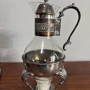 May include: A vintage glass and silver coffee pot with a decorative handle and base. The pot features a clear glass carafe with a silver metal band and lid. The base has ornate, curved legs.