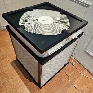 May include: An air purifier with a black frame and a white fan on top. The filter is visible below the fan, and the unit sits on a wooden floor. The air purifier is designed to clean the air in a room.