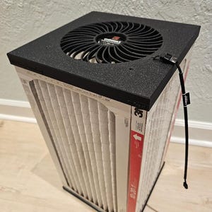 May include: An air purifier with a black top and fan, and a white filter. The filter has a red stripe with text and an arrow. A black cord extends from the top.