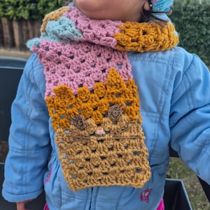 May include: A handmade crochet scarf in shades of pink, gold, and blue. The scarf features a cat face design with brown whiskers and nose. The scarf is worn over a blue jacket.