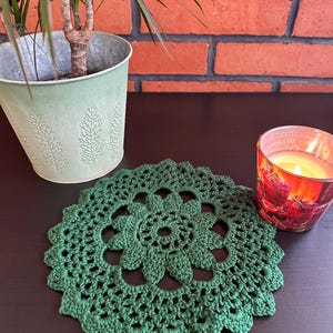 Può includere: Un centrino all'uncinetto verde fatto a mano con un motivo floreale. Il centrino è circolare e si trova su una superficie scura. Una candela accesa in un barattolo di vetro e una pianta in vaso sono sullo sfondo.