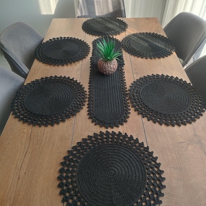 Peut inclure: Une table à manger en bois avec six sets de table en crochet noirs et un chemin de table assorti. Une petite plante décorative dans un pot marron est posée sur le chemin de table. Les sets de table sont ronds avec un bord décoratif.