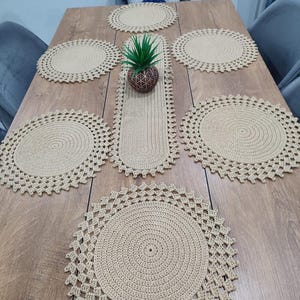 Peut inclure: Un ensemble de table en crochet beige, comprenant six dessous de plats ronds et un chemin de table rectangulaire. Une petite plante en pot est au centre. Les dessous de plats et le chemin de table ont un motif ajouré détaillé.