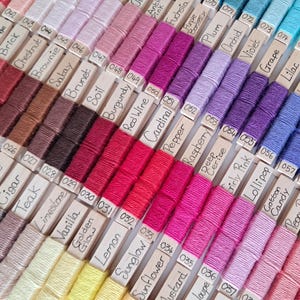 May include: A close-up of a colour palette of embroidery floss, arranged in rows. The floss is wound on small wooden bobbins, each labelled with a colour name. Colours range from reds and pinks to purples and blues.