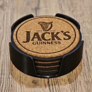 May include: A set of round cork coasters with the text "Jack's Guinness" and a harp logo, held in a black holder. The coasters are made of natural cork material and are designed to protect surfaces from drink condensation.