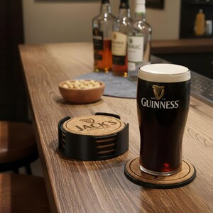 May include: A pint of Guinness stout with a white head sits on a cork coaster. A set of cork coasters with the text "Jack's Guinness" is next to the pint. A small bowl of peanuts and bottles are in the background.