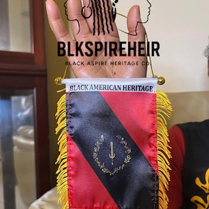 Puede incluir: Una pequeña bandera rectangular con un diseño rojo y negro, adornada con flecos dorados. La bandera dice "BLACK AMERICAN HERITAGE" y presenta un emblema dorado. La marca "BLKSPIREHEIR" es visible encima de la bandera.