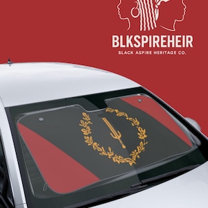 Puede incluir: Parasol para coche con un diseño negro y rojo, con un emblema dorado y el texto "BLKSPIREHEIR BLACK ASPIRE HERITAGE CO." El parasol se muestra en un coche, ofreciendo protección solar.